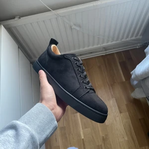 Louboutin skor - Stilrena svarta sneakers i mocka från Christian Louboutin med klassisk röd sula och svarta snören. Skorna har rund tå, platt sula och diskreta sömdetaljer. Perfekta för dig som vill ha en lyxig och clean look med en ikonisk touch.