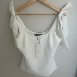 Vit topp med rosetter från Zara - Aldrig använt!! Trendig vit topp från Zara med breda axelband som knyts i stora rosetter på axlarna. Toppen har figurnära passform och en stilren, elegant look. Slut i S på hemsidan ❤️ 