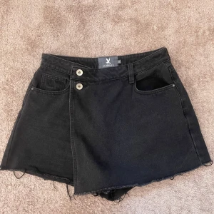 Svarta jeansshorts från CC Double O - Svarta jeansshorts från CC Double O med råa kanter och cool omlott-look framtill. Shortsen har hög midja, två knappar och dragkedja. Klassisk femficksmodell i denim som ger en edgy vibe. Perfekta för dig som gillar streetstyle och vill sticka ut lite extra.
