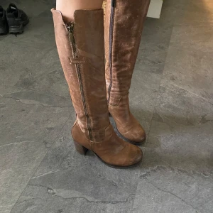 Bruna höga skinnstövlar med klack i vintage stil - Snygga bruna stövlar i skinn med hög skaft och markerad klack. Dragkedja på sidan ger en cool detalj och gör dem enkla att ta på. Rund tå och robust sula, perfekta till jeans eller kjol för en trendig look.