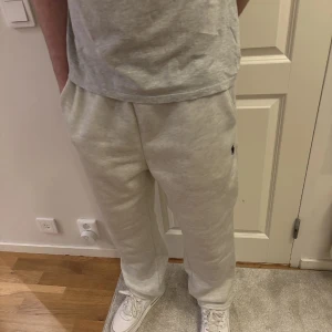 Grå mjukisbyxor från Polo Ralph Lauren - Snygga ljusgrå mjukisbyxor från Polo Ralph Lauren med elastisk midja och sidofickor. Byxorna har en liten svart broderad logga på ena benet och är tillverkade i mjuk bomull. Perfekta för en avslappnad stil. Passform: Baggy/loose