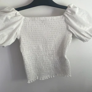 Vit smockad topp med puffärm Gina Tricot - Supersöt vit topp från Gina Tricot i smockat tyg med bred resår över hela kroppen och stora puffärmar. Offshoulder-modell som ger en trendig vibe. Säljer då den inte används storlek xs