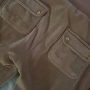 Beiga mjukisbyxor Juicy Couture S - Supermysiga beiga mjukisbyxor från Juicy Couture med stora fickor framtill och snygga guldfärgade knappar. Byxorna har dragsko i midjan för perfekt passform och är gjorda i mjukt material som känns lyxigt mot huden.