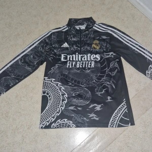 Real Madrid x Adidas draktröja svart  - Snygg långärmad Real Madrid fotbollströja från Adidas i svart med grått drakmönster. Tröjan har dragkedja vid halsen, vita ränder på ärmarna och klubbmärke i guld. Materialet är lätt och andas, perfekt för träning eller match.