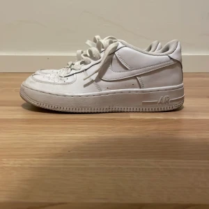 Nike Air Force 1 vita sneakers - Klassiska Nike Air Force 1 sneakers i helvitt skinn med rund tå och platt sula. Snygg och tidlös modell med snörning och diskret Nike-logga på sidan och hälen. Perfekta för dig som gillar streetstyle och vill ha en clean look.