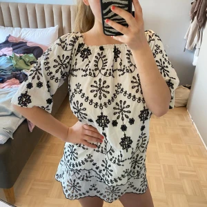Vit kortklänning med svart broderi - Supersöt vit kortklänning med svart broderat blommönster och scallop-kant. Offshoulder-modell med resår upptill och vida trekvartsärmar. Perfekt för sommarens alla tillfällen, luftig och lätt i bomull.