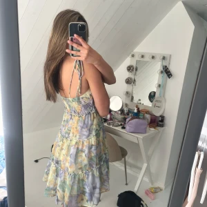 blommig volangklänning - Supersöt kortklänning med blommigt pastellmönster i lila, gul och grön. Klänningen har tunna axelband som knyts, volanger i flera lager och smockad rygg. Perfekt för sommarens alla tillfällen och ger en riktigt fräsch vibe.