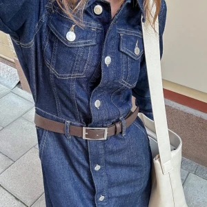 Maxi denim klänning - Säljer en lång mörkblå jeansklänning med klassiska silverfärgade knappar, bröstfickor. Klänningen har lång ärm, markerad midja och raka linjer för en cool och tidlös look. Perfekt för dig som gillar denim och vill sticka ut.