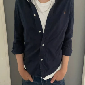 Mörkblå skjorta från Ralph Lauren - Snygg mörkblå skjorta från Ralph Lauren med klassisk krage och vita knappar. Skjortan har lång ärm och den ikoniska broderade loggan på bröstet. Perfekt att bära öppen över en t-shirt för en avslappnad stil.