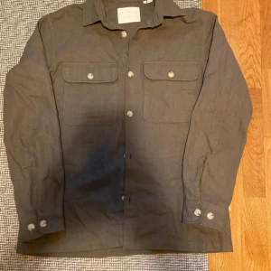 Brun overshirt från Jack & Jones M - Snygg mörkgrön overshirt från Jack & Jones med klassisk krage och två stora bröstfickor med knappar. Skjortan är långärmad och har knäppning framtill samt vid ärmsluten. Perfekt att bära som lager över en t-shirt.