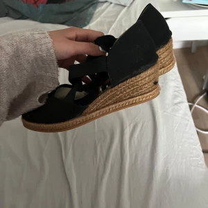 Svarta kilklack-sandaler från Dasia - Snygga svarta sandaler med kilklack från Dasia. Ovandel i tyg med elastiska band som korsas över foten för en cool look. Klacken är täckt med flätad jute för en somrig vibe. Perfekta till jeans eller klänning när du vill ha lite höjd men ändå vara bekväm.