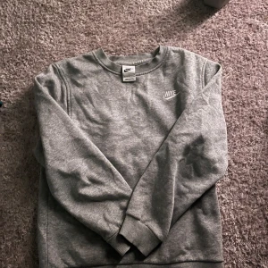 Grå crewneck från Nike - En stilren grå crewneck från Nike med broderad logga på bröstet. Tröjan har rund halsringning, långa ärmar och ribbade muddar. Perfekt för en chill och sportig look. Materialet är mjukt och skönt, troligtvis bomullsmix.