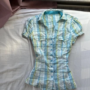 Rutig kortärmad skjorta i blått och grönt - Kortärmad skjorta med rutigt mönster i blått, grönt och vitt. Skjortan har figurnära passform, krage, knappar framtill och två bröstfickor med lock. Materialet är lätt och känns som bomull, perfekt för varmare dagar.