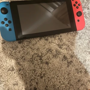 Kommer med fodral och laddare . Brukade spela förr men inte längre och funkar galant  - Bilderna visar en Nintendo Switch-konsol med blå och röd Joy-Con, tillhörande dockningsstation och ett svart fodral. Perfekt för gaming på språng eller hemma! Inga böcker syns på bilderna.