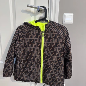 Fendi vindjacka med FF-mönster - Snygg vindjacka från Fendi med ikoniskt FF-mönster i brunt och svart. Jackan har huva med neonfoder, dragkedja i neonfärg och två fickor framtill. Perfekt för dig som vill sticka ut med en lyxig streetstyle-look.