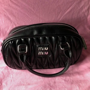 Svart quiltad handväska Miu Miu - Säljer en svart quiltad handväska i skinn med silverfärgade detaljer och logotyp framtill. Väskan har både handtag och en avtagbar axelrem, perfekt för att bära på olika sätt. Stilren oval form och dragkedja upptill.