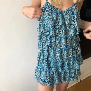 Blå mönstrad volangklänning - Supersöt kort blå klänning med tunna axelband och flera lager volanger. Klänningen har ett paisleymönster i blått, svart och beige, samt öppen rygg. Perfekt för sommarens varma dagar och ger en riktigt fräsch vibe. står ej storlek men skulle gissa s/m! pris kan diskuteras