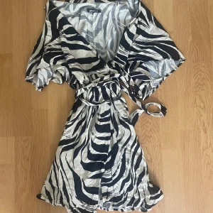 Zebramönstrad omlottklänning Gina Tricot - Snygg kort omlottklänning från Gina Tricot i zebramönster. Klänningen har svartvita ränder, korta ärmar och knyts i midjan med ett bälte. Perfekt för dig som vill sticka ut med en trendig look.