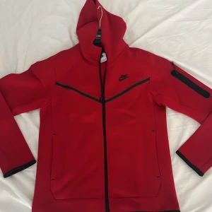 Röd Nike tech fleece-kofta - Säljer en röd Nike tech fleece-kofta endast använd en gång för 800kr pris kan diskuteras.