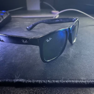Svarta Ray-Ban Wayfarer solglasögon - Klassiska svarta Ray-Ban Wayfarer solglasögon med fyrkantig form och mörka glas. Snygg och tidlös design med Ray-Ban-logga på båda sidor av bågen. Perfekta för dig som vill ha en ikonisk look och skydda ögonen mot solen.