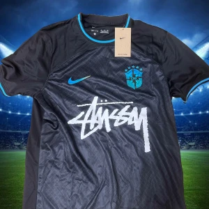 fotbollströja Brasilien x Stüssy  - Snygg svart fotbollströja från Nike i samarbete med Stüssy och Brasilien. Tröjan har korta ärmar, blå detaljer vid krage och ärmslut, samt blå Nike-logga och Brasilien-emblem. Stüssy-loggan är tryckt i vitt framtill. Tillverkad i lätt och ventilerande material.