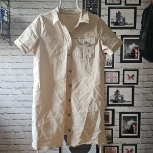 Skjortklänning från VILA - Stilren beige overshirt från VILA i kraftig bomull. Skjortan har korta ärmar, klassisk krage, knappar framtill och en bröstficka med knapp. Perfekt att bära öppen eller stängd för en avslappnad look.