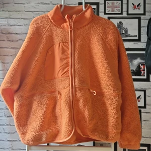 Orange teddyjacka med dragkedja - Säljer en orange teddyjacka med hög krage och hel dragkedja framtill. Jackan har två stora fickor med dragkedja och en bröstficka med knapp. Perfekt för dig som gillar en chill och cozy stil. Materialet är mjukt fleece-liknande och jackan har en loose passform.