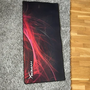 Hyperx musmatta - Bilderna visar en stor musmatta från HyperX med svart bakgrund och röda grafiska mönster. Produkten är perfekt för gaming eller datorarbete, 
