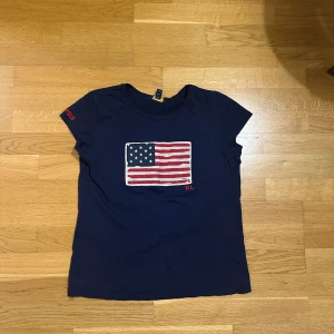 Mörkblå t-shirt från Polo Ralph Lauren - Barnstorlek 176 (16 år) Mörkblå t-shirt från Polo Ralph Lauren med amerikansk flagga broderad på bröstet och RL-detalj. Kortärmad modell i mjuk bomull, perfekt för en avslappnad och cool stil. Polo-tryck på ärmen ger extra edge.