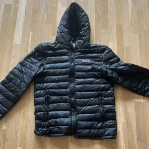 Svart pufferjacka från Patagonia - Säljer en svart pufferjacka från Patagonia med huva och dragkedja framtill. Jackan är quiltad och har två fickor med dragkedja. Tillverkad i 100% polyester och har en clean, sportig look som funkar till allt. Perfekt för kyliga dagar!