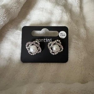 Pärlörhängen med strass från Montini - Snygga örhängen från Montini med en stor vit pärla i mitten och glittrande strass runtom i en blomformad design. Perfekta för att ge din look lite extra bling och elegans. Fästs med klassisk plupp på baksidan.