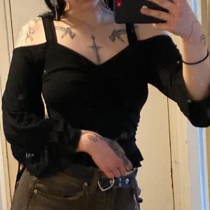 Svart offshoulder topp med volang - Säljer en svart offshoulder topp med långa ärmar och breda axelband. Toppen har volanger nertill och vid ärmslut, samt snygg rynkdetalj framtill. Croppad modell som ger en edgy vibe, perfekt att styla med höga jeans. Från Gina tricot
