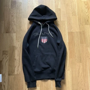 Svart hoodie från GANT med brodyr - Svart hoodie från GANT med broderad logga och sköld på bröstet. Tröjan har huva med vita snören, känguruficka och ribbade muddar. Perfekt för dig som gillar klassisk och sportig stil. Materialet är mjuk bomullsmix.