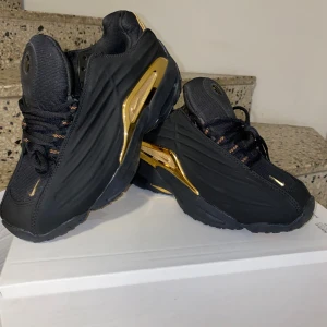 Svarta Nocta sneakers med guldiga detaljer - Snygga svarta sneakers med coola guldiga detaljer på sidorna och sulan. Skorna har snörning och en sportig vibe, med gulddetaljer som verkligen sticker ut. Yttersulan har inslag av gult och ovandelen är i textil och syntet. Perfekta för dig som vill ha något unikt på fötterna.