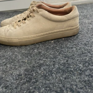 Beige mockasneakers från Polo Ralph Lauren - Snygga beige sneakers från Polo Ralph Lauren i mocka med diskret logga bak och på sulan. Klassisk låg modell med snörning och platt sula. Perfekta för en clean och stilren look.