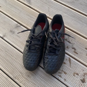 Nike Tiempo svarta fotbollsskor - Säljer ett par Nike Tiempo fotbollsskor i svart med röda detaljer och snörning. Skorna har en quiltad ovandel i syntetmaterial och rund tå. Sulan har röda dobbar för bra grepp på planen. Perfekta för dig som vill sticka ut på matchen!