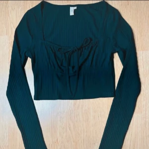 Svart croppad topp från Nelly - Svart långärmad crop top från Nelly med ribbad struktur, fyrkantig urringning och knytning framtill – en feminin och smickrande modell. Stretchigt material i återvunnet tyg för bekväm passform. Storlek S (tight men bekväm). Toppskick. Mått: längd 41 cm, byst 82 cm, midja 68 cm. Material: 64 % återvunnen polyester, 33 % viskos, 3 % elastan. Prutning ej möjlig – men jag erbjuder rabatt vid köp av flera plagg. Rensar garderoben – kika gärna på mina andra annonser!  ⸻  