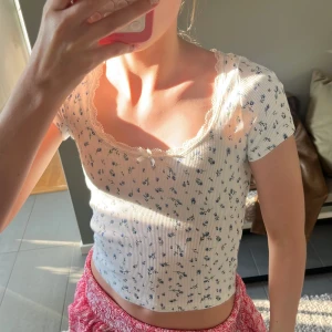 Vit blommig croppad topp med spets - Supersöt croppad topp i vit ribbad bomull med små blå blommor. Toppen har korta ärmar, spetskant vid urringningen och en liten rosett framtill. Perfekt för en somrig look och passar till både jeans och kjol.