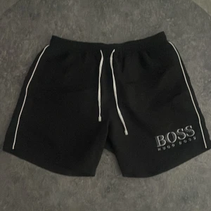 Svarta badshorts från Hugo Boss - Svarta badshorts från Hugo Boss med vit snörning i midjan och vita detaljer längs sidorna. Stort BOSS-tryck på ena benet. 