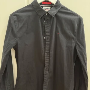 Svart skjorta Tommy Jeans slim fit M - Snygg svart skjorta från Tommy Jeans i slim fit-modell. Skjortan har klassisk krage, knäppning framtill och diskret logga på bröstet. 
