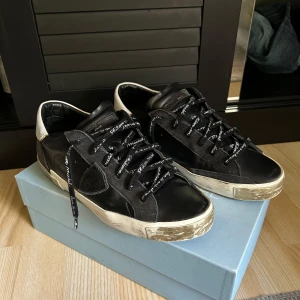 Philippe Model Skor  - Svarta Philippe Model sneakers med vita detaljer och logga på hälen. Skorna har snörning med svarta skosnören med vit text, platt sula och rund tå. Ovandelen är i skinn och mocka, med slitna detaljer på skon, det slitna är med meningen. Modellen är så. Skick: 8,5/10. Sulan är väldigt bra för att vara ett par Philippe model skor. ÄKTA SÅ KLART. För fler frågor så är det bara att ställa dom.
