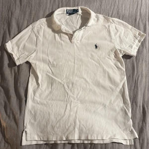 Vit pikétröja från Polo Ralph Lauren - Klassisk vit pikétröja från Polo Ralph Lauren med korta ärmar och den ikoniska broderade loggan på bröstet. Tröjan har krage och knäppning vid halsen. Tillverkad i mjuk bomull som är skön att bära hela dagen.