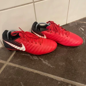 Röda Nike fotbollsskor med dobbar - Snygga röda fotbollsskor från Nike med svarta och vita detaljer. Skorna har snörning och klassiska dobbar för bra grepp på planen. Ovandelen är i syntetmaterial och designen är sportig med en stor vit swoosh på sidan.