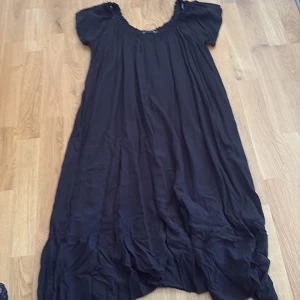 Svart långklänning från 157 - Svart långklänning med lös passform från 157. Klänningen har korta ärmar, rundad och rynkad halsringning samt ett luftigt fall. Perfekt för dig som gillar en enkel och avslappnad stil. Materialet känns lätt och svalt.