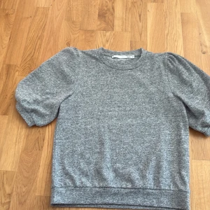 Grå kortärmad tröja från 157 - Mysig grå tröja från 157 med korta puffärmar och rund halsringning. Tröjan har en enkel och stilren design med mjukt material som känns skönt mot huden. Perfekt för lager-på-lager eller bara som den är till jeans eller kjol.