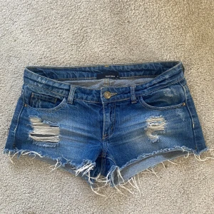 Blå slitna jeansshorts Tally Weijl - Snygga blå jeansshorts från Tally Weijl med slitna detaljer och fransig kant. Klassisk femficksmodell med låg midja och dragkedja. Perfekta för sommaren och ger en cool, avslappnad vibe. Små i storleken!