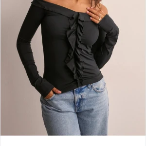 Svart offshoulder topp med volang - Säljer en svart offshoulder topp med långa ärmar och snygg volangdetalj framtill. Toppen har en tight passform och är perfekt för dig som gillar en stilren men ändå trendig look. Materialet känns mjukt och stretchigt mot huden.