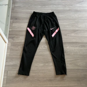 Svarta PSG träningsbyxor från Nike - Svarta träningsbyxor från Nike med PSG-logga och rosa detaljer på framsidan. Byxorna har smal passform, elastisk midja och dragkedja vid benslut. Perfekta för fotboll eller träning och tillverkade i ett lätt syntetmaterial.