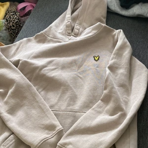 Beige hoodie från Lyle & Scott - Snygg beige hoodie från Lyle & Scott med klassisk gul broderad logga på bröstet. Tröjan har huva med breda snören och en stor magficka. Mjuk bomullskänsla och långärmad modell, perfekt för chill dagar.
