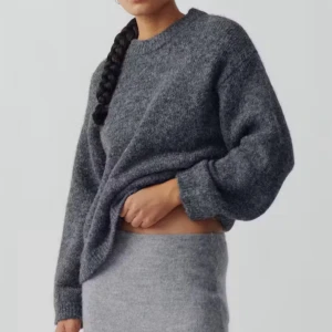 Grå stickad tröja med rund hals - Mysig grå stickad tröja med rund hals och långa ärmar. Tröjan har en avslappnad passform och ribbade muddar vid ärmslut och nederkant. Perfekt för kyliga dagar och enkel att matcha med jeans eller kjol.